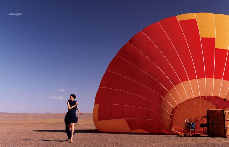 Model Qun Ye walks across an arid desert terrain beside a large red and orange hot air balloon envelope under a clear blue sky.