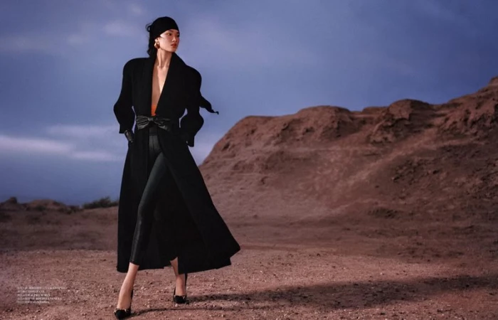 Model Qun Ye posing in a long black belted coat, black headscarf, and heels against a desert hill landscape at dusk.