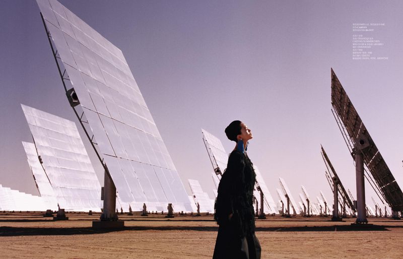 Model Qun Ye posing in a black textured coat among rows of towering solar heliostat panels in a desert landscape.