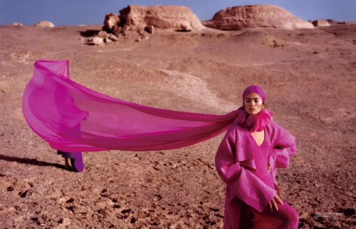 Model Qun Ye posing in a vibrant magenta coat with a dramatic flowing veil across an arid desert landscape.