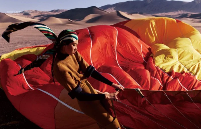 Model Qun Ye stands in a desert holding rigging ropes of a giant red and yellow hot air balloon canopy.