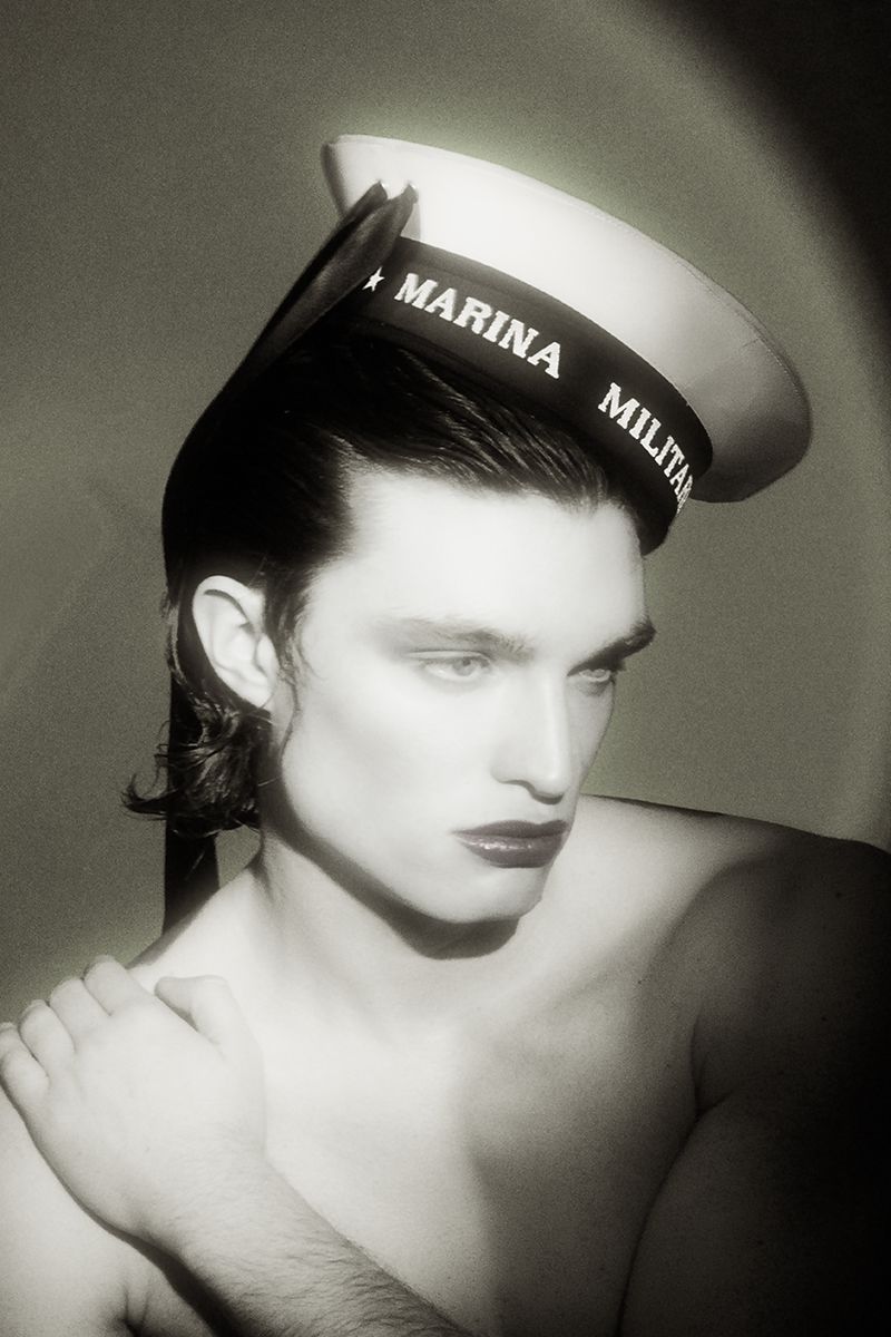 A shirtless man wearing an Italian naval sailor hat tilted on his head, looking to the side with dark lipstick in spotlight lighting.