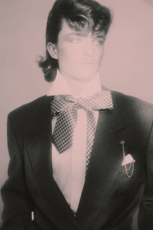 Model with retro wavy hairstyle wearing a dark tailored blazer and oversized houndstooth bow tie against a soft studio backdrop.