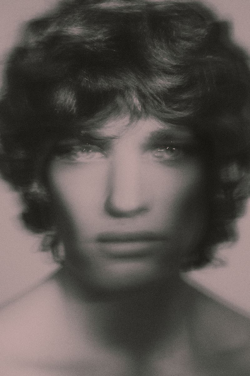 Soft-focus close-up portrait of a young man with dark wavy hair looking directly forward against a muted backdrop.