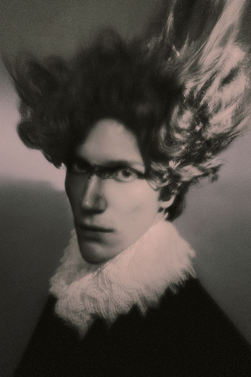 Portrait of a man with dramatic swept-back hair wearing a white ruffled collar and dark makeup across his eyes.