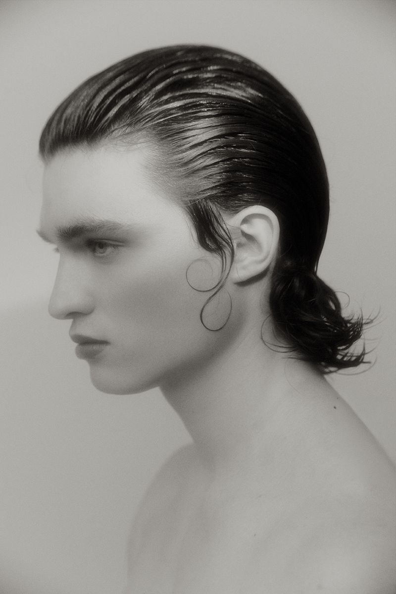 Monochrome profile portrait of a young man with slicked-back dark hair and a curled sideburn strand against a seamless light backdrop.