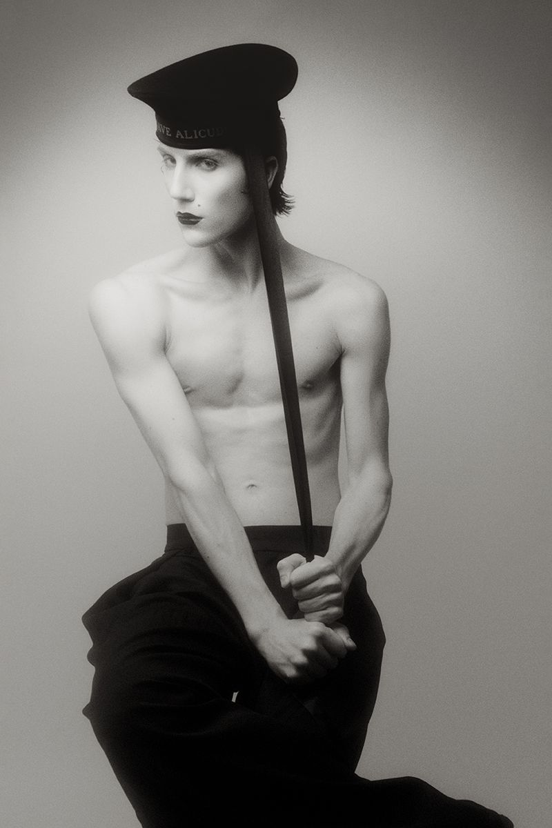 A shirtless man wearing dark lipstick and a black sailor hat poses while holding a strap against a grey studio backdrop.