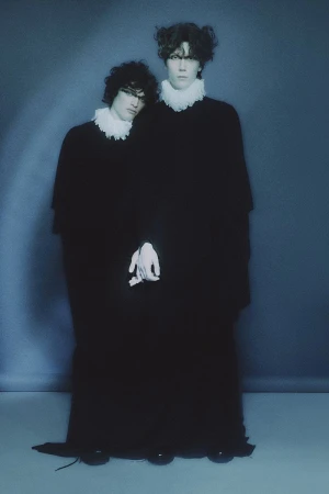 Two young men with dark curly hair wearing voluminous black robes and white ruffled collars against a moody blue background.