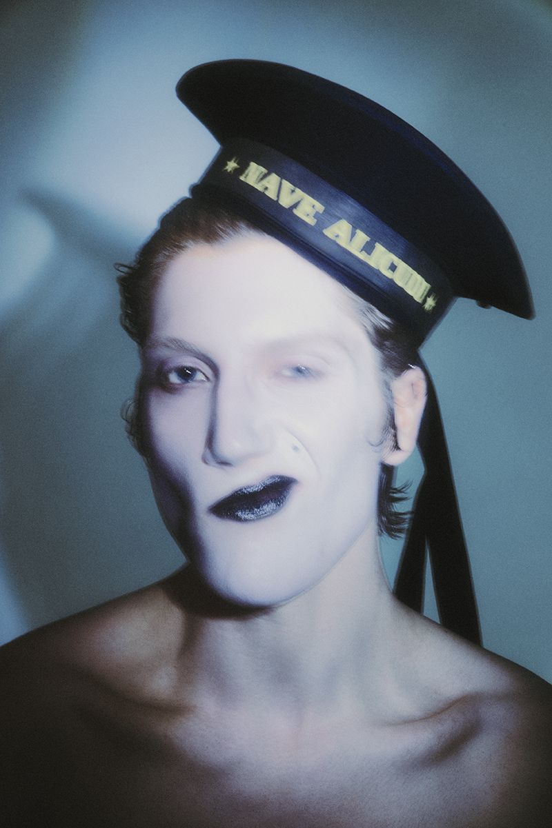 Model with dramatic white face makeup and dark lips wearing a black sailor hat against a cool-toned studio background.
