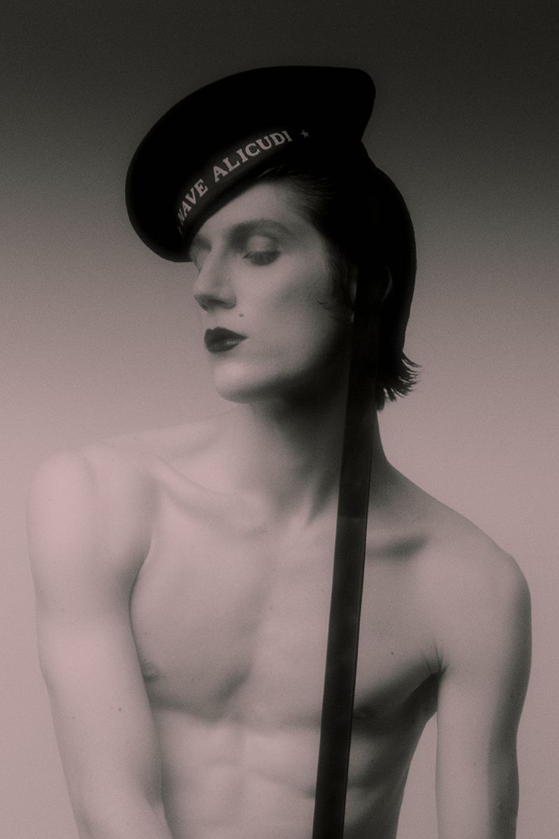 Shirtless young man wearing dark lipstick and a black naval cap inscribed with Nave Alicudi in a studio portrait.