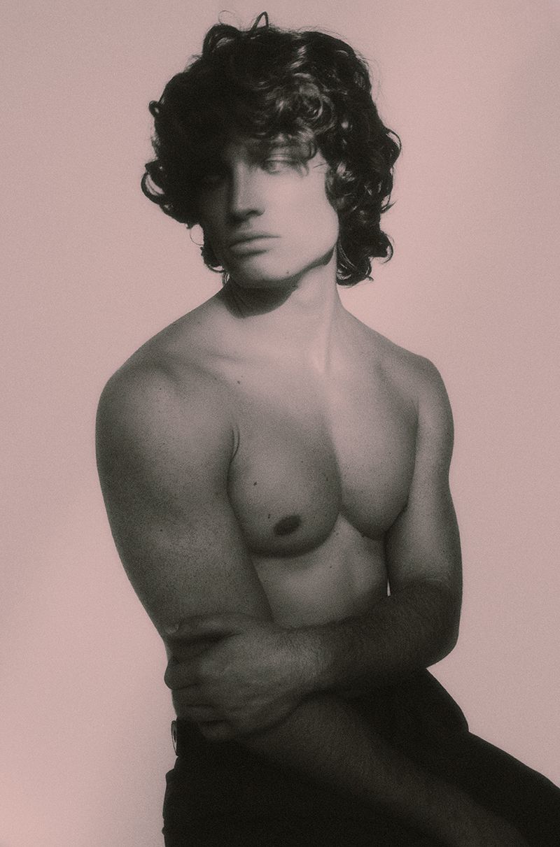 Shirtless young man with dark curly hair posing in dramatic soft lighting against a muted pale studio backdrop.