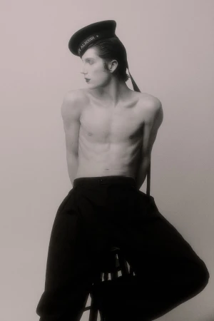 Shirtless young man wearing a black sailor hat and high-waisted black trousers seated against a pale studio backdrop.