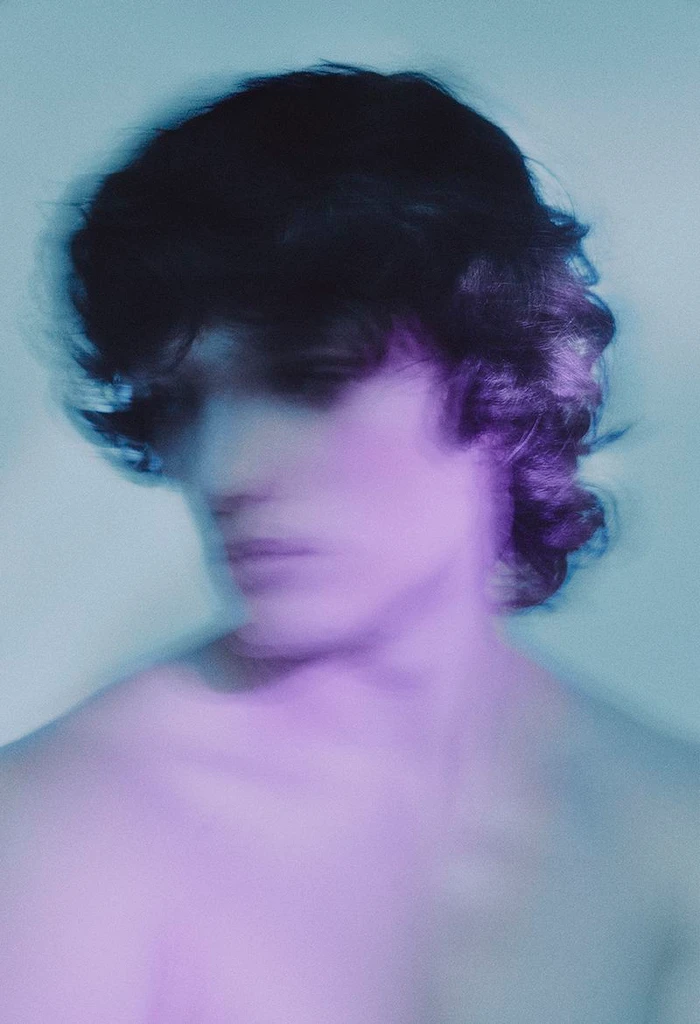Blurred portrait of a shirtless young man with dark wavy hair illuminated by soft purple and cyan lighting.