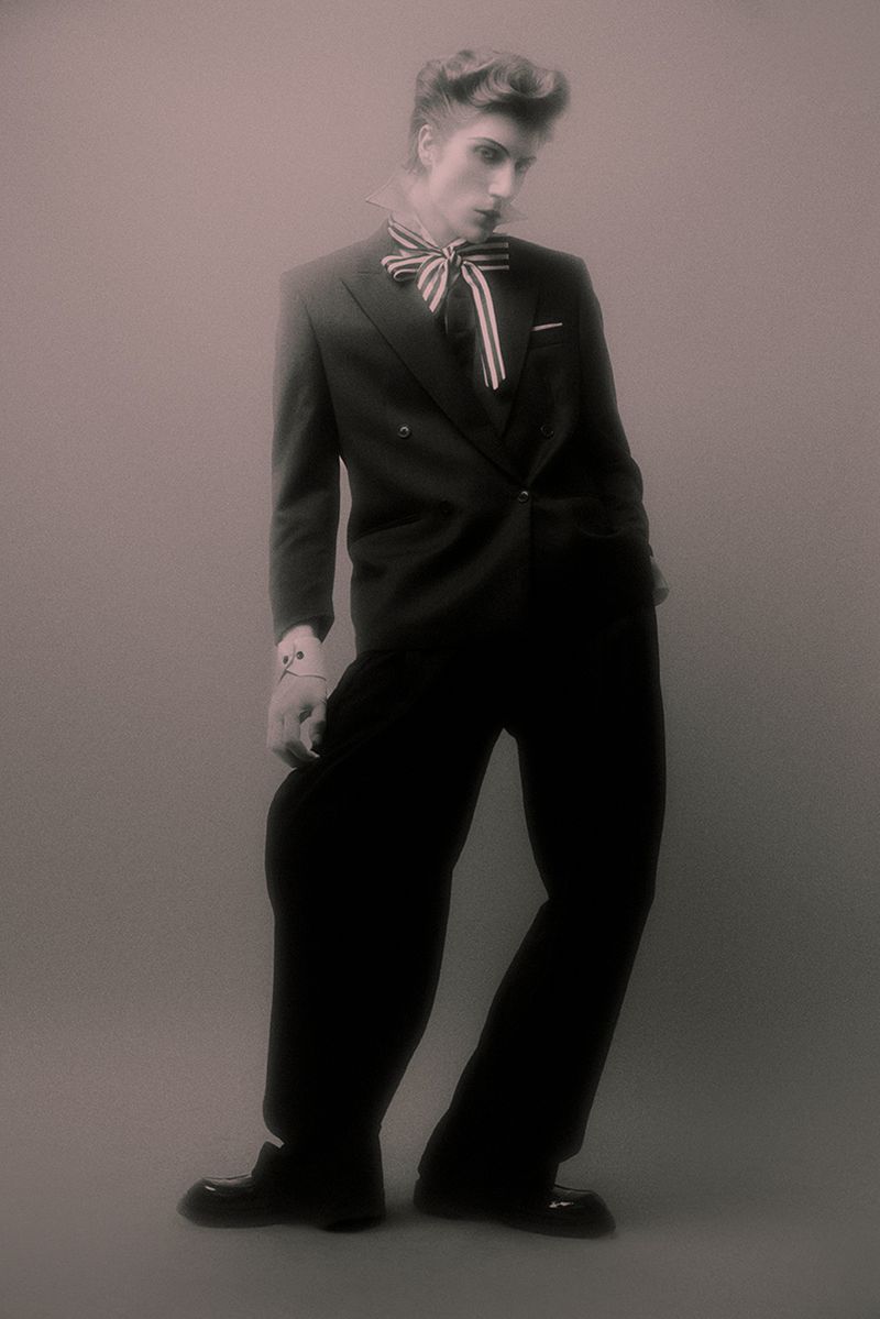 Model in a black double-breasted suit and striped neck ribbon posing against a grey backdrop with a vintage quiff hairstyle.