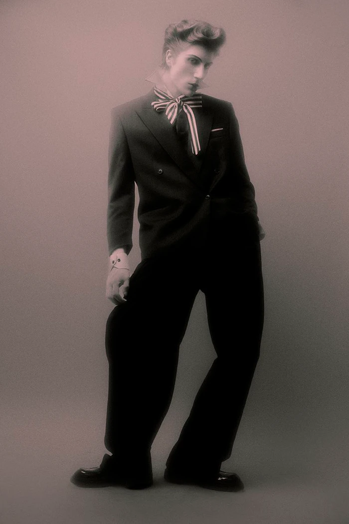 Model in a black double-breasted suit and striped neck ribbon posing against a grey backdrop with a vintage quiff hairstyle.