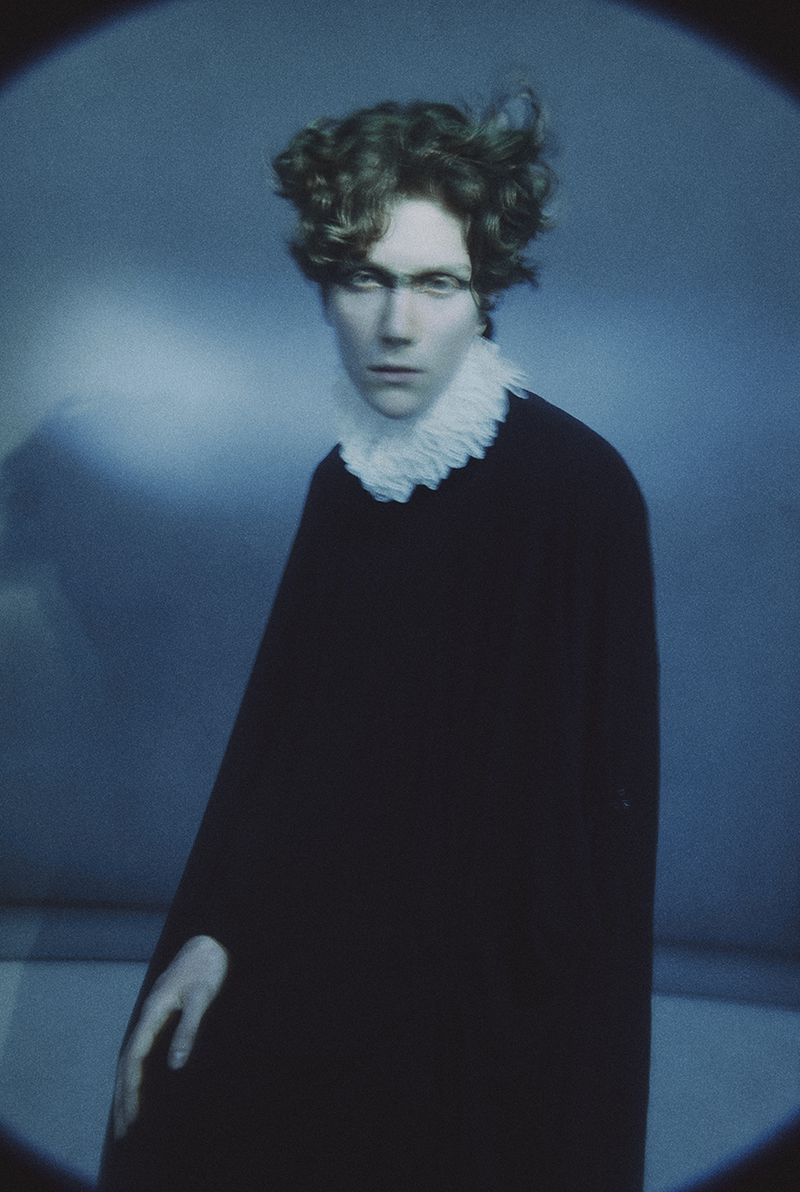 Editorial portrait of a person with styled wavy hair wearing a black robe and white ruffled collar against a cool blue background.
