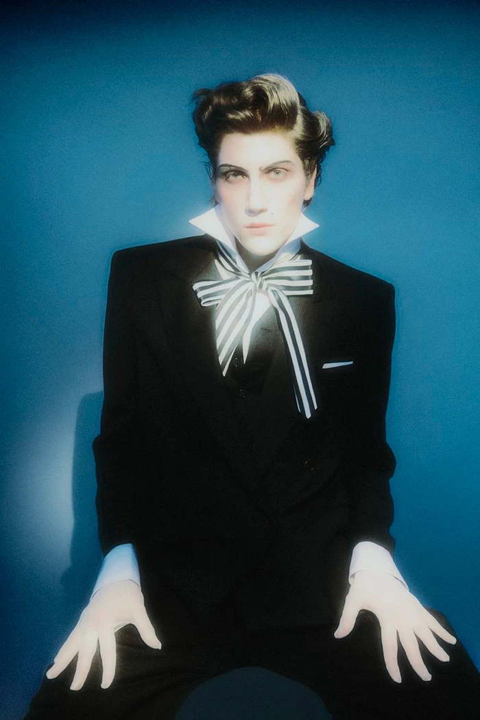 Fashion portrait of a model in a black suit with a large striped bow tie and theatrical makeup against a blue backdrop.