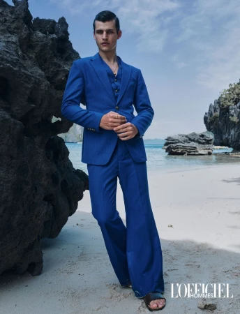 Male model in a vibrant blue three-piece suit standing on a sandy beach against dark coastal rocks.