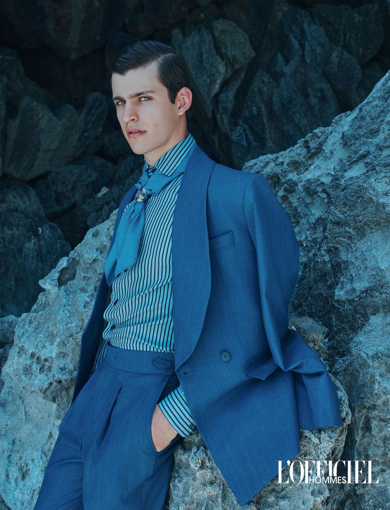 Model wearing a tailored blue suit and striped shirt with a neck scarf, leaning against rocky coastal cliffs.