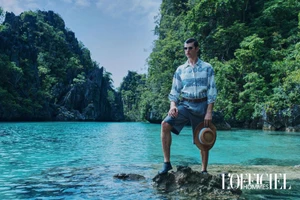 Male model in a patterned shirt and grey shorts standing on rocks in turquoise lagoon water holding a straw hat.