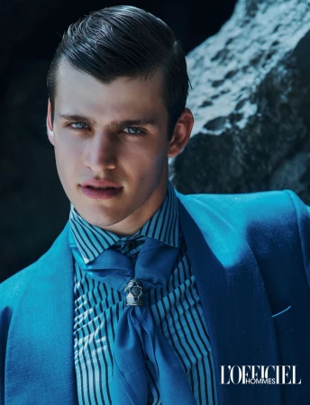 A male model with slicked dark hair wearing a bright blue suit, striped shirt, and blue ascot in front of a rocky background.