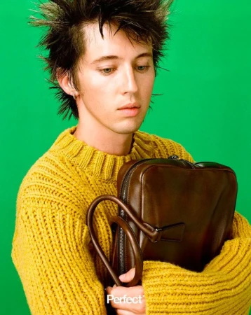 Troye Sivan holding a brown leather bag while wearing a yellow ribbed knit sweater against a green studio backdrop.