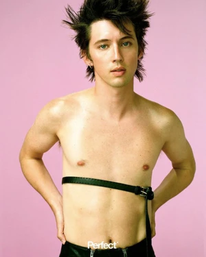 Troye Sivan posing shirtless with spiky hair and a black leather chest strap against a pink background.