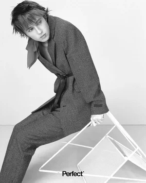 Troye Sivan wearing a belted textured suit seated on a white geometric chair against a light background for The Perfect Magazine.