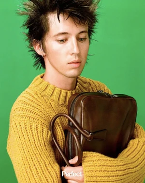 Troye Sivan with spiky hair in a yellow ribbed knit sweater holding a brown leather bag against a green backdrop.