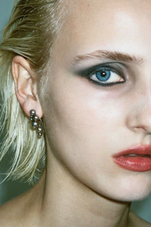 Close-up of a blonde model with dark smokey eye makeup, red lips, and a silver beaded earring in an editorial portrait.