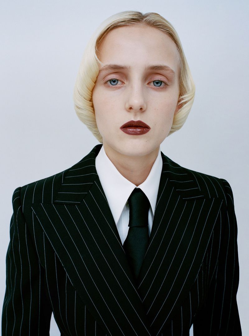 Model in a black pinstripe suit, white collared shirt, and black tie posing with dark lipstick against a white studio background.