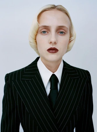Model in a black pinstripe suit, white collared shirt, and black tie posing with dark lipstick against a white studio background.