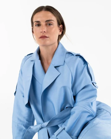 Headshot of a fair-skinned adult woman looking at the camera, wearing a light blue trench coat against a bright white background.