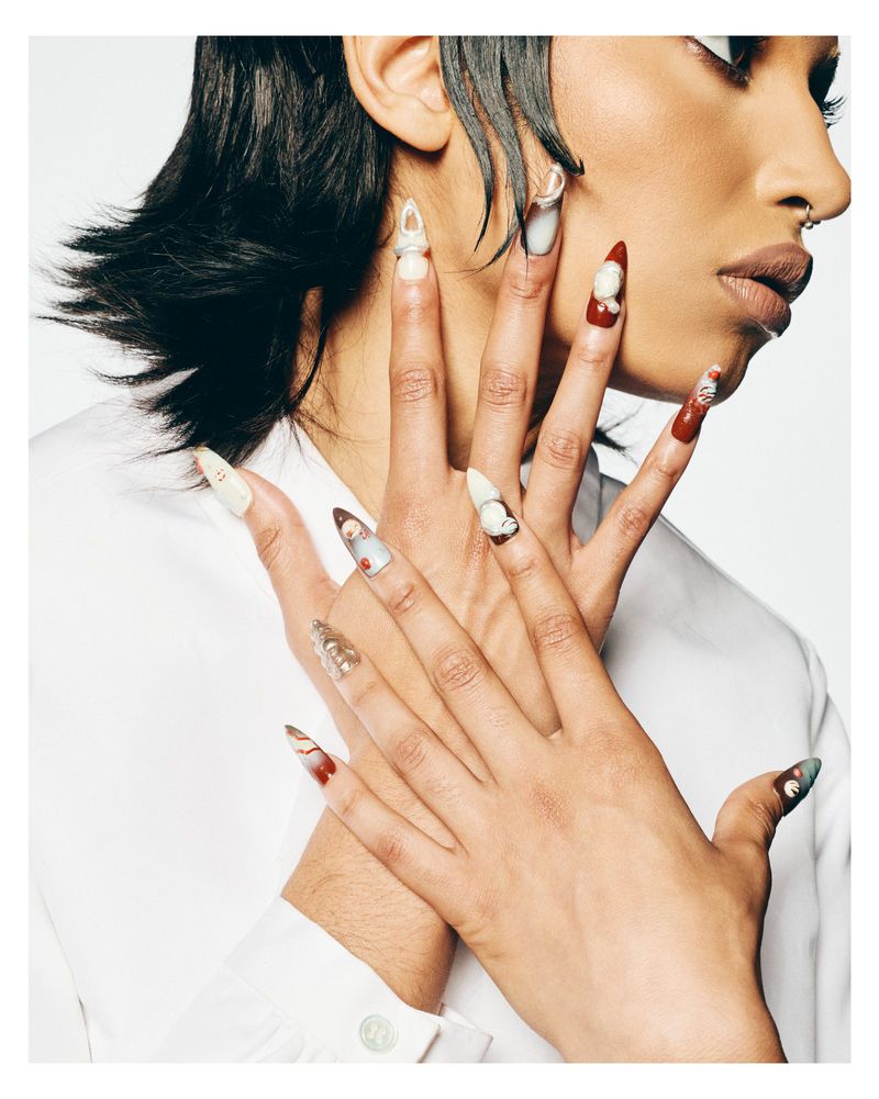 Close-up beauty portrait featuring hands with elaborate sculpted 3D nail art posed against the neck and face, wearing a white shirt.