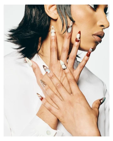 Close-up beauty portrait featuring hands with elaborate sculpted 3D nail art posed against the neck and face, wearing a white shirt.