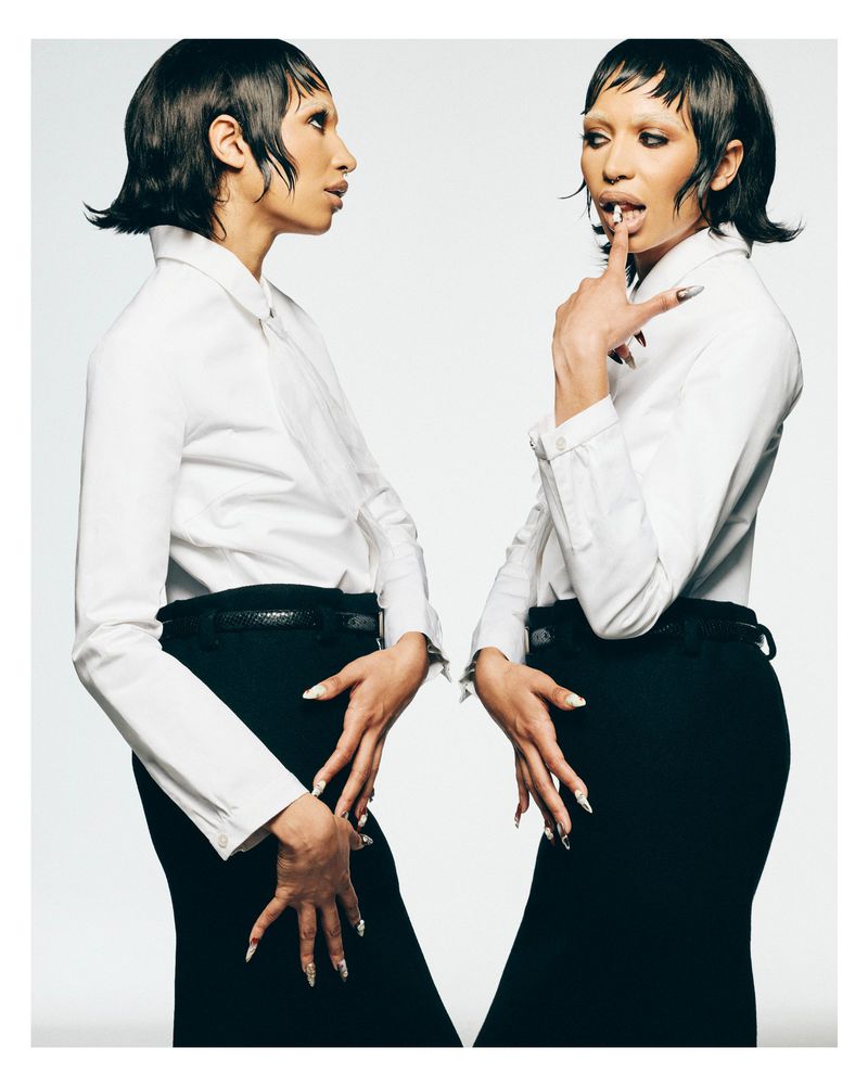 Model in a white collared shirt and black skirt with short micro-bang hair against a seamless white studio backdrop.