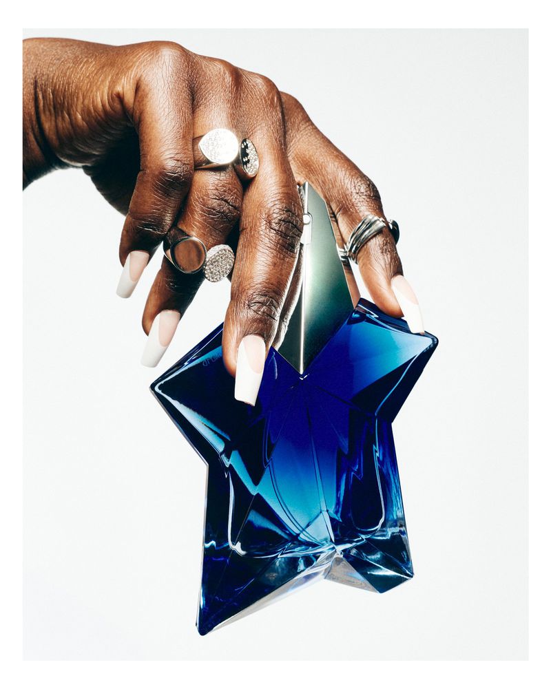 A hand with dark skin and silver rings holds a faceted blue star-shaped perfume bottle against a solid white background.