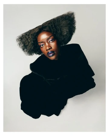 High-angle beauty portrait of a Black woman with sculptural flared hair, glossy dark lips, and a black garment against a white backdrop.