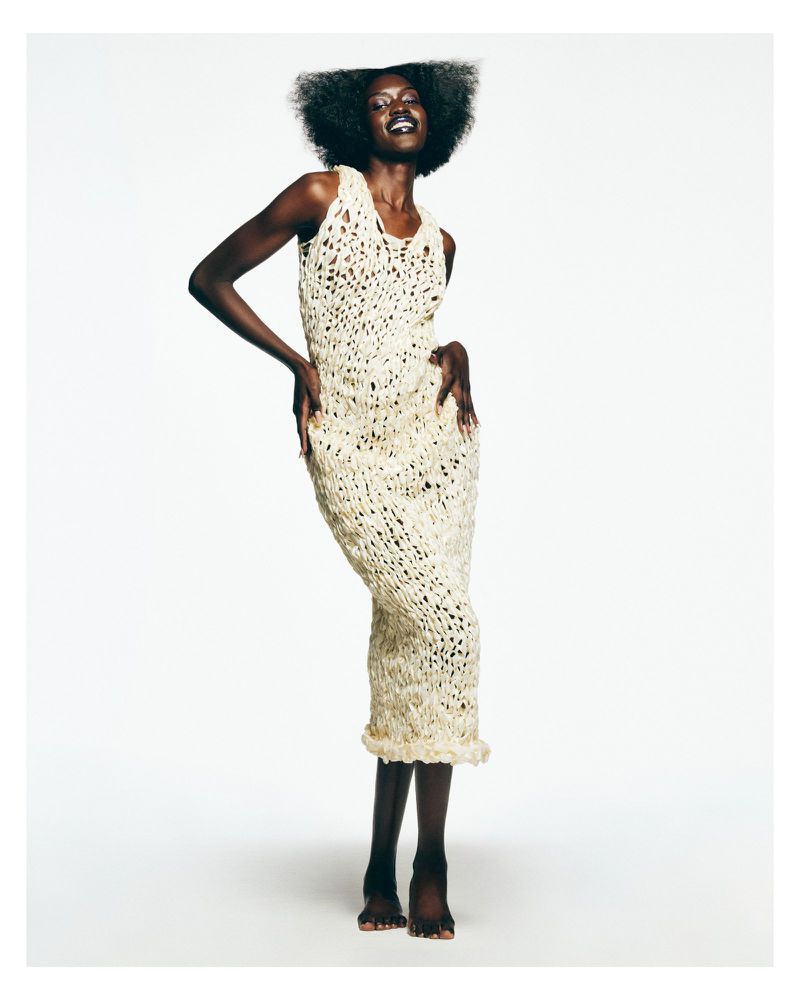 A smiling Black woman posing barefoot in a sleeveless cream crochet midi dress against a seamless white studio background.