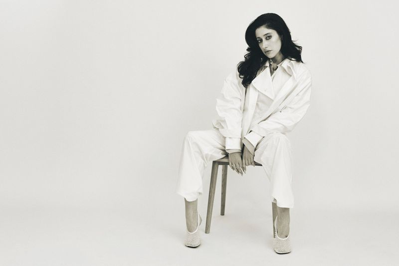 Taylor Ortega wearing an oversized white suit seated on a wooden chair against a white seamless studio background.