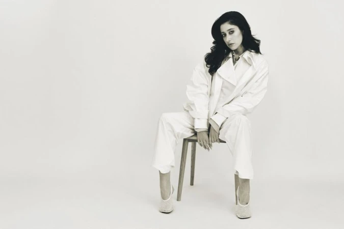 Taylor Ortega wearing an oversized white suit seated on a wooden chair against a white seamless studio background.