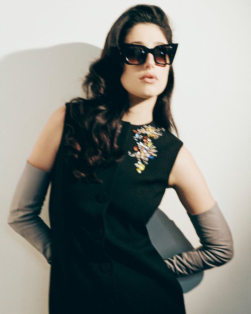 Taylor Ortega wearing black cat-eye sunglasses, a black embellished sleeveless dress, and grey opera gloves against a white background.