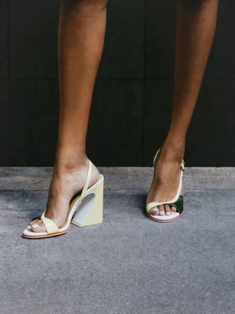 Close-up of model Selina Pillay wearing pale yellow sculptural high-heel sandals while standing on a concrete ground.