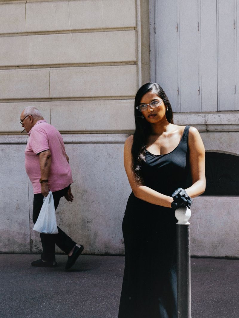 Model Selina Pillay in a black sleeveless dress and glasses leaning on a bollard with a pedestrian walking past behind her.