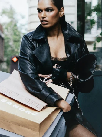 Model Selina Pillay wearing a black leather trench coat and holding paper shopping bags on a city street.