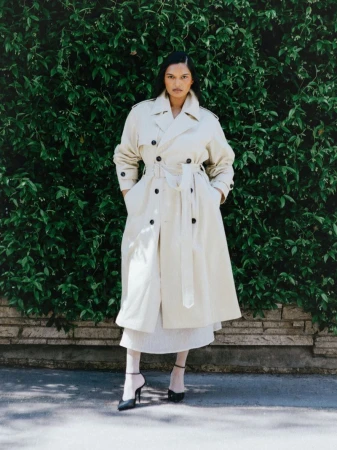 Model Selina Pillay in a belted beige double-breasted trench coat and black heels standing before a green foliage hedge.