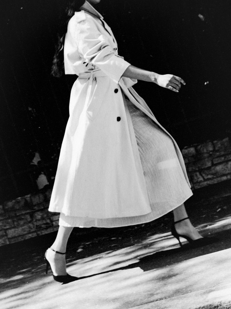Selina Pillay walking in a light belted trench coat and black ankle-strap heels in black and white.