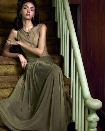 A woman in a sheer olive green sleeveless floor-length gown seated on carpeted indoor stairs beside a pale green railing.