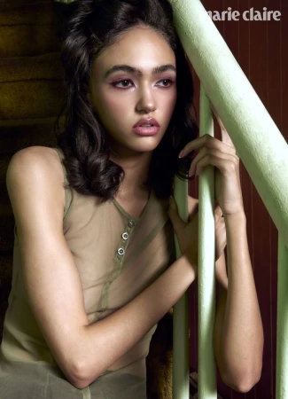 Model with dark wavy hair wearing a sheer sleeveless top seated next to a pale green stair railing.