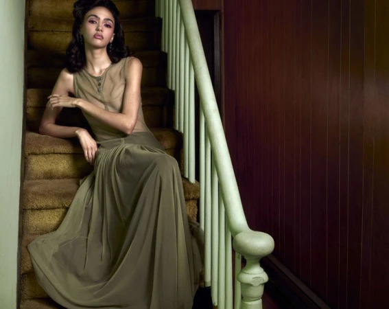 A female model in a draped olive green gown seated on a carpeted staircase beside a painted wooden railing.
